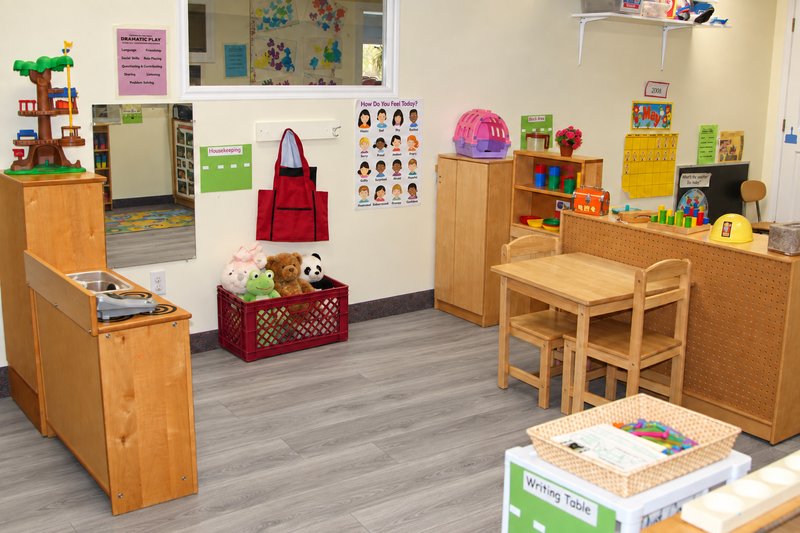 Auburn dramatic-play area with a wooden play kitchen, dress-up apron, stuffed animals, and child-sized table