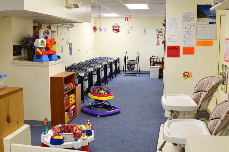 Auburn infant room with a carpeted play area, infant activity toys, and feeding chairs